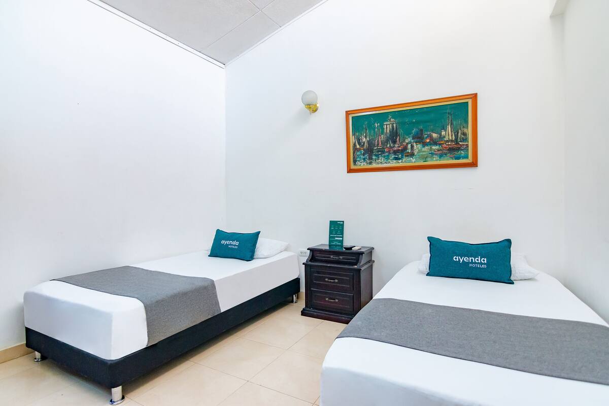 Habitación de hotel con dos camas, cada una con cubrecama gris y blanco, y un almohadón color turquesa con el nombre del hotel. En la pared, un cuadro que representa un paisaje urbano.