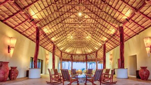 A spacious indoor area with a high ceiling and a thatched roof structure. There are several rocking chairs arranged around a central table, and large vases are placed on the floor.