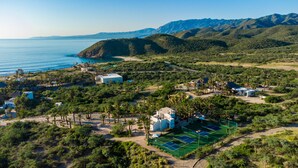 Luftaufnahme eines Küstenresorts mit mehreren Gebäuden, einem Tennisplatz und einem Strandbereich.