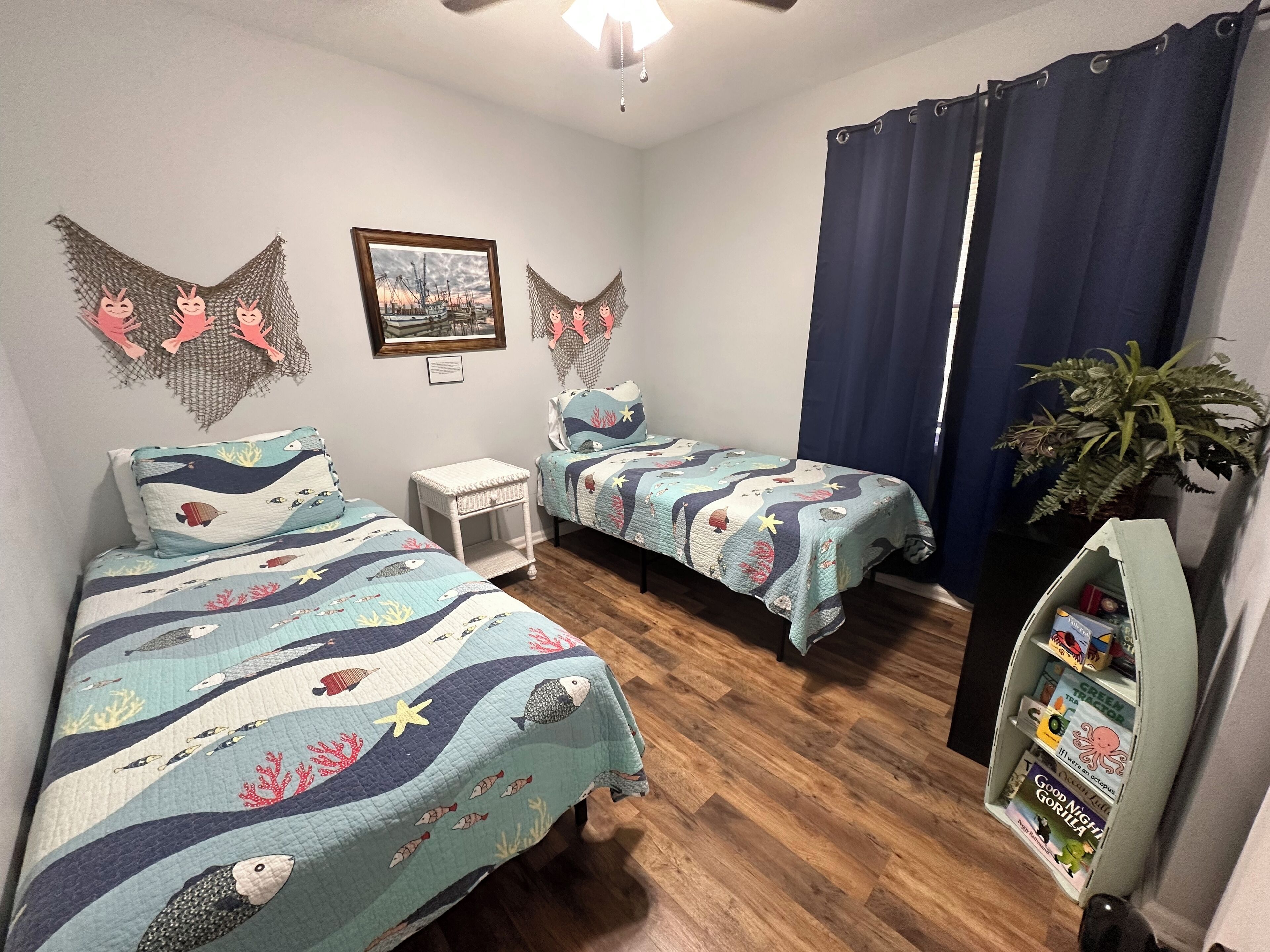 A room with two beds, a small table, a bookshelf, and a ceiling fan.