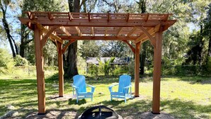 Una pérgola de madera con dos sillones Adirondack azules y un brasero en un jardín trasero cubierto de césped.