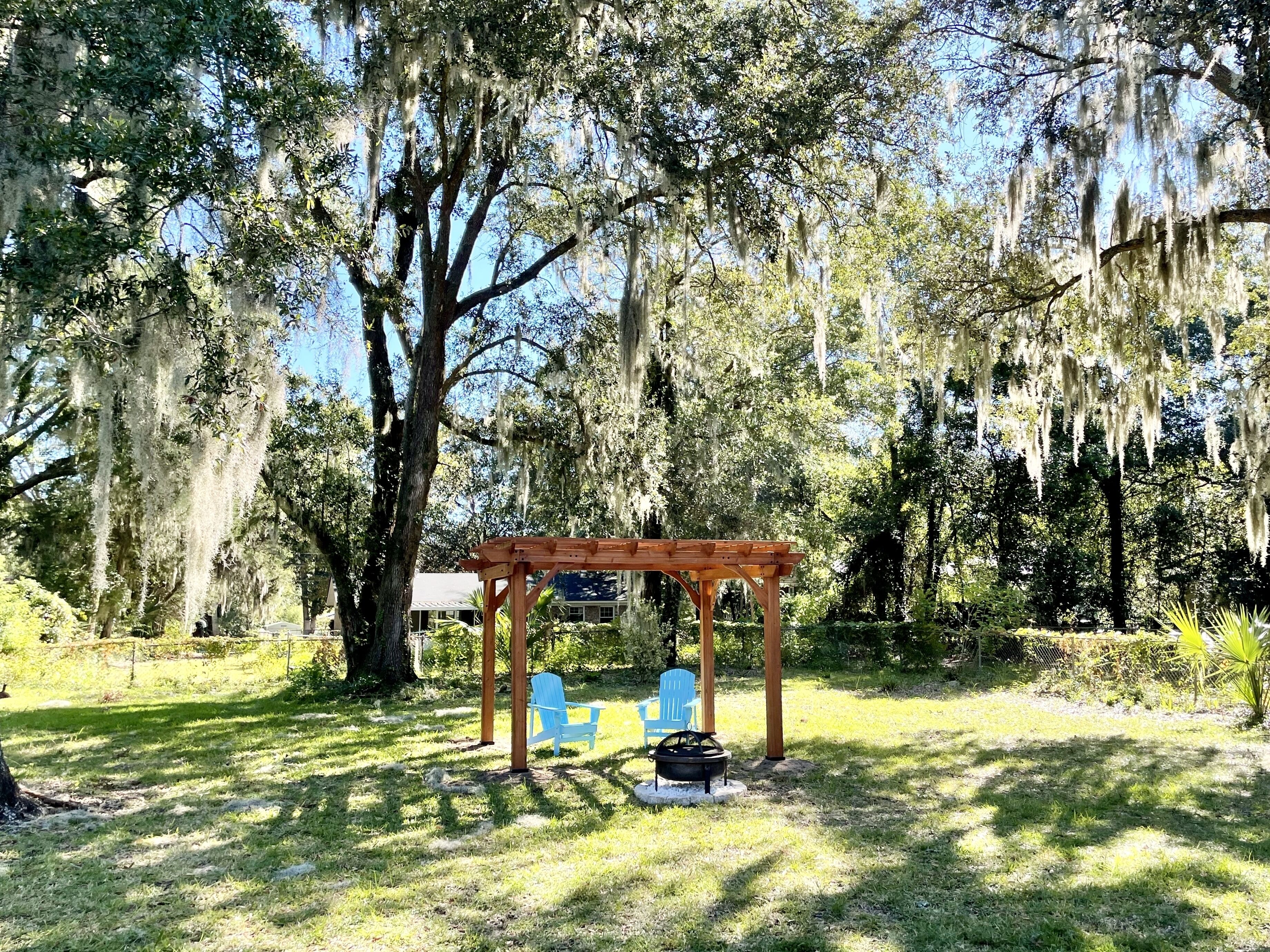 A wooden pergola with a fire pit and two blue chairs in a grassy outdoor area with trees and Spanish moss.