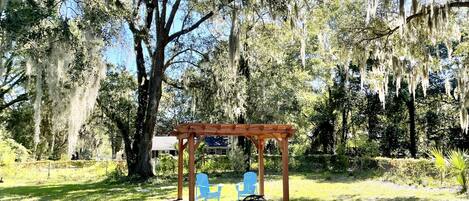 A wooden pergola with a fire pit and two blue chairs in a grassy outdoor area with trees and Spanish moss.