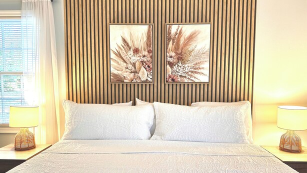 Habitación de hotel con cama, dos almohadas, cabecero de madera con listones verticales, dos cuadros florales colgados en la pared y lámparas de noche.