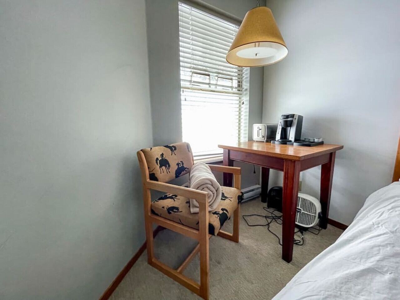 Habitación de hotel pequeña con una cama individual, una silla de madera con cojín estampado, un escritorio con lámpara y una ventana con persianas.