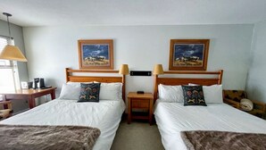 A hotel room with two single beds, a desk with a chair, a lamp, and two framed pictures on the wall. - Apex Mountain ~ Hillside Hideout ~ Apex Mountain Inn (Apex Mountain Resort)