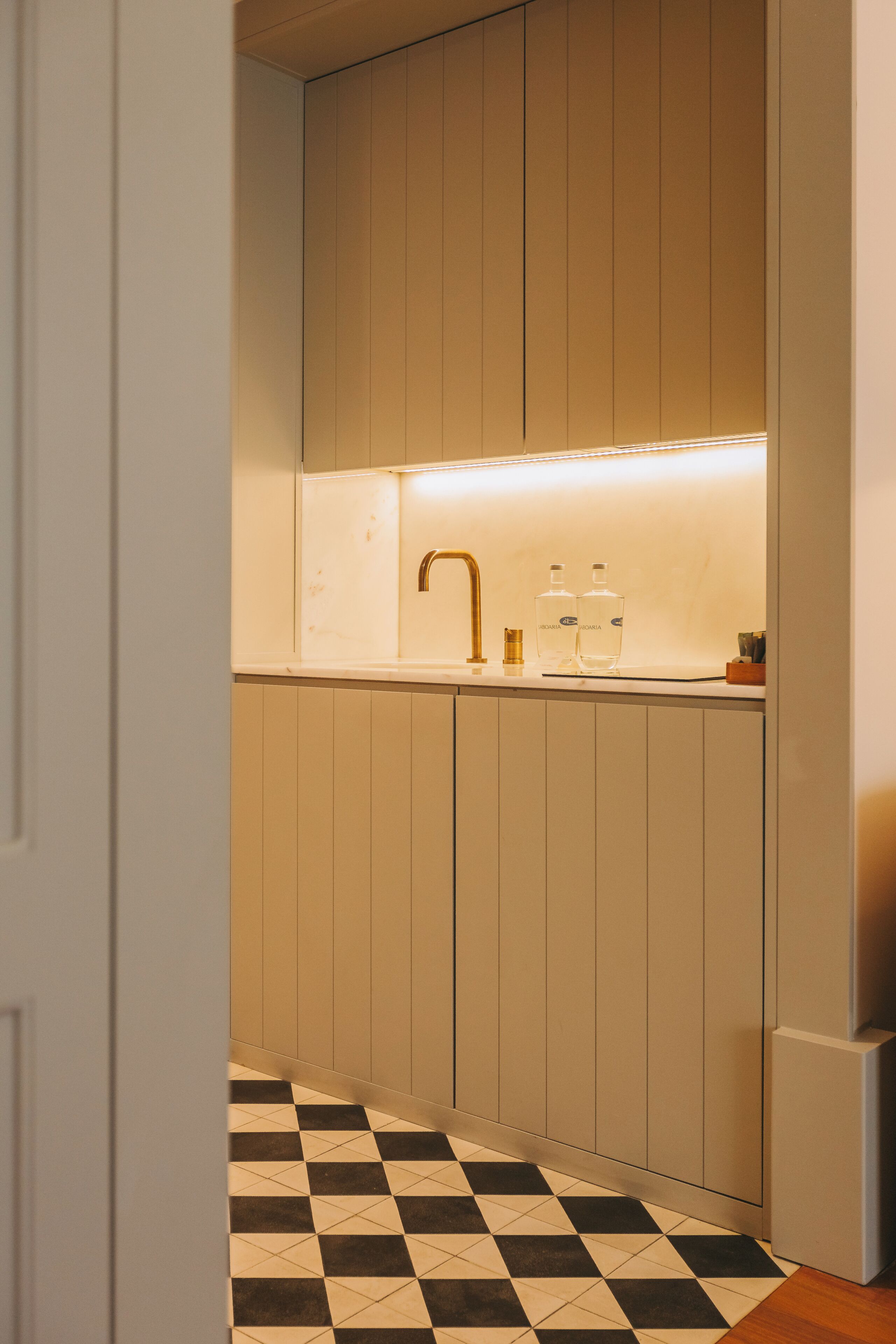 A narrow hallway with a checkered floor leading to a kitchen area with a sink, faucet, and two bottles on the counter.