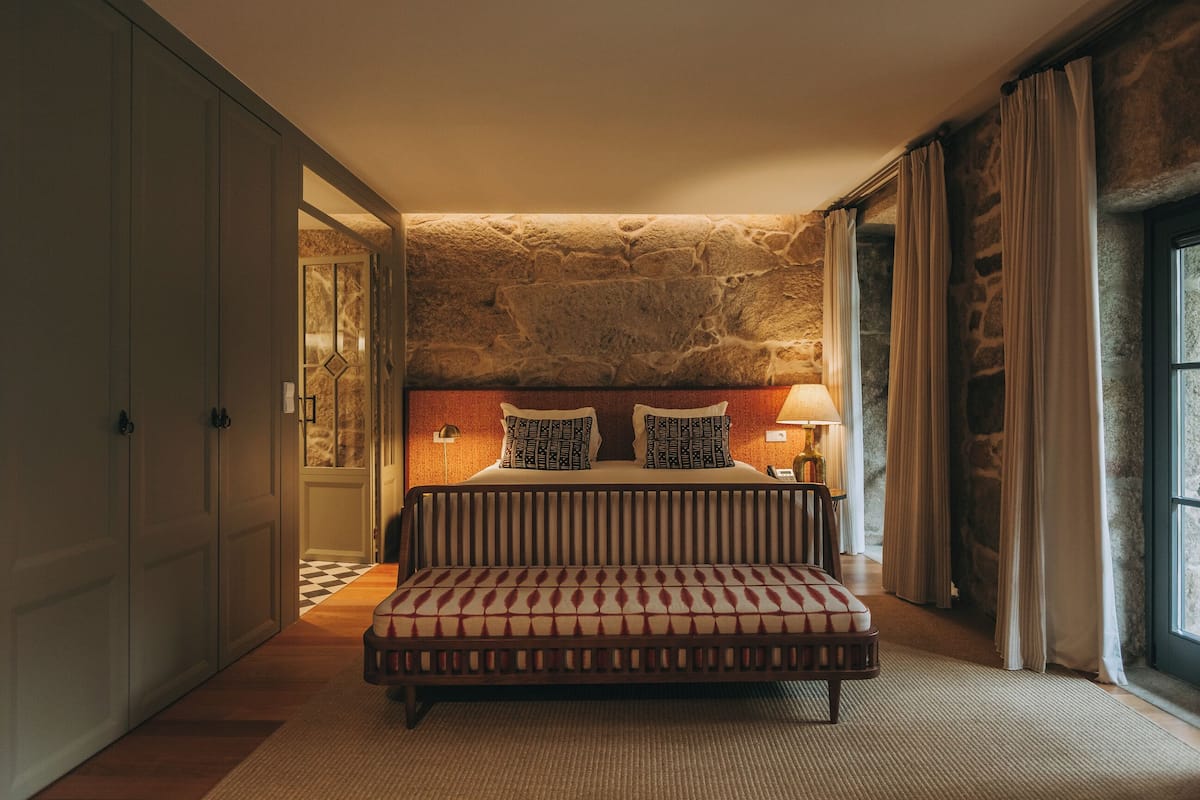 A bedroom with a stone wall, a bed with patterned pillows, a bench, and a nightstand with a lamp.