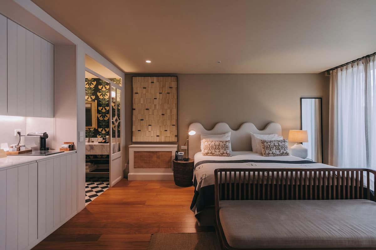 A modern hotel room with a bed, bedside lamps, a wooden headboard, a small table, and a bench.