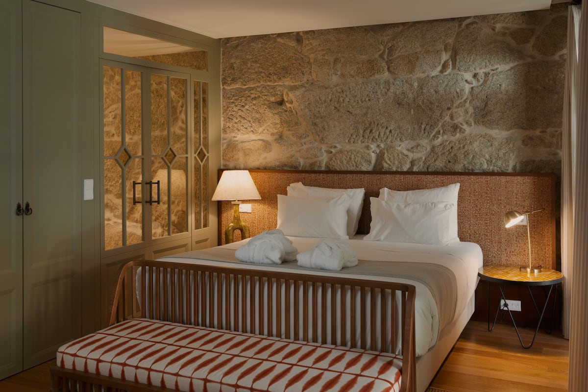 A hotel room with a large bed, a wooden bench, a stone wall, and a nightstand with a lamp.