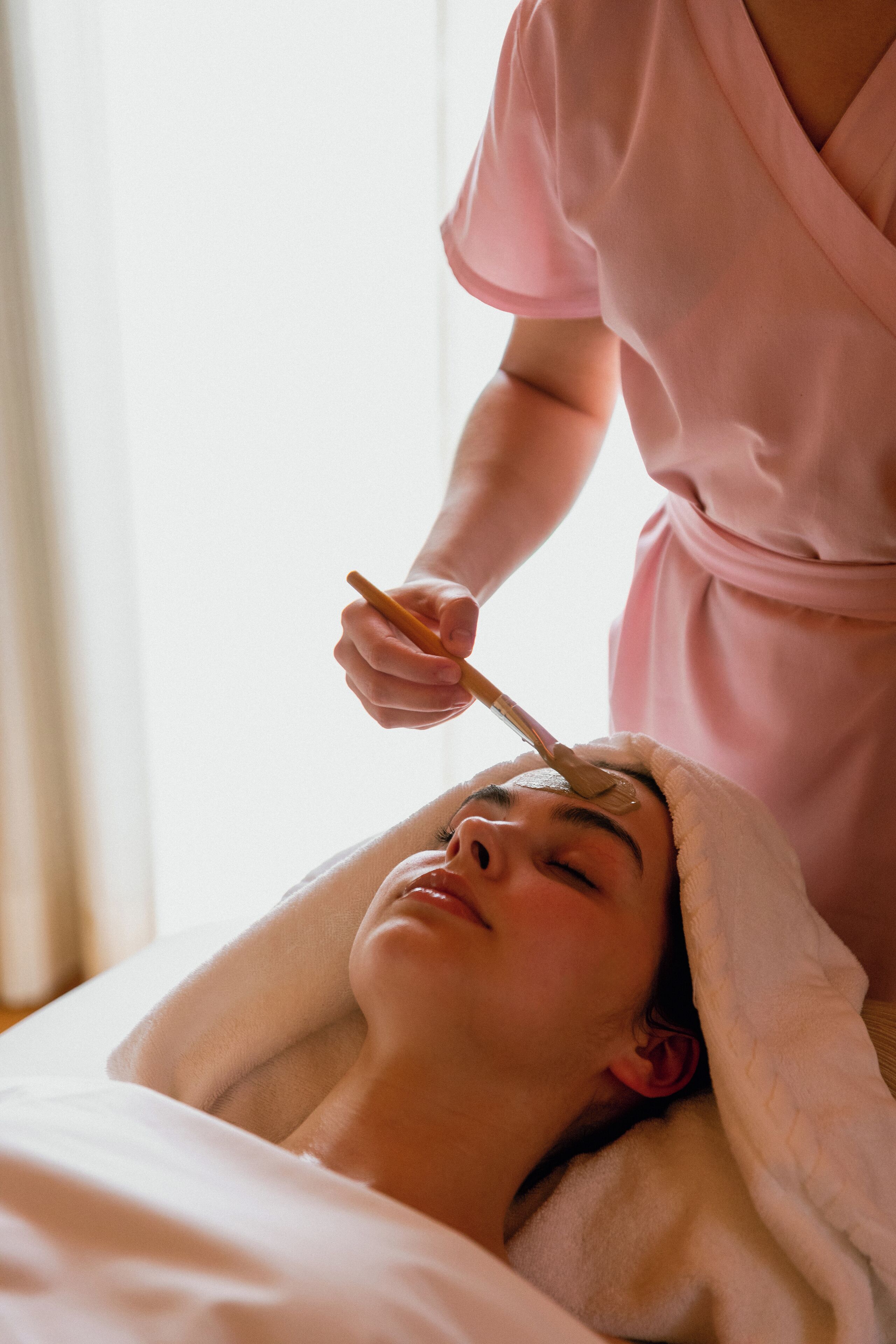 Una persona recibe un tratamiento facial en una habitación con una cama y un ventanal.