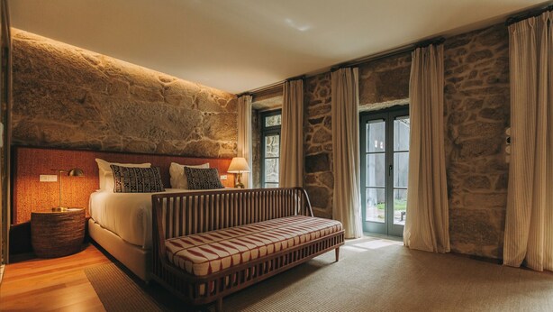 A bedroom with a stone wall, a bed, a bench, and a large window with curtains. - Torel Saboaria (Porto)