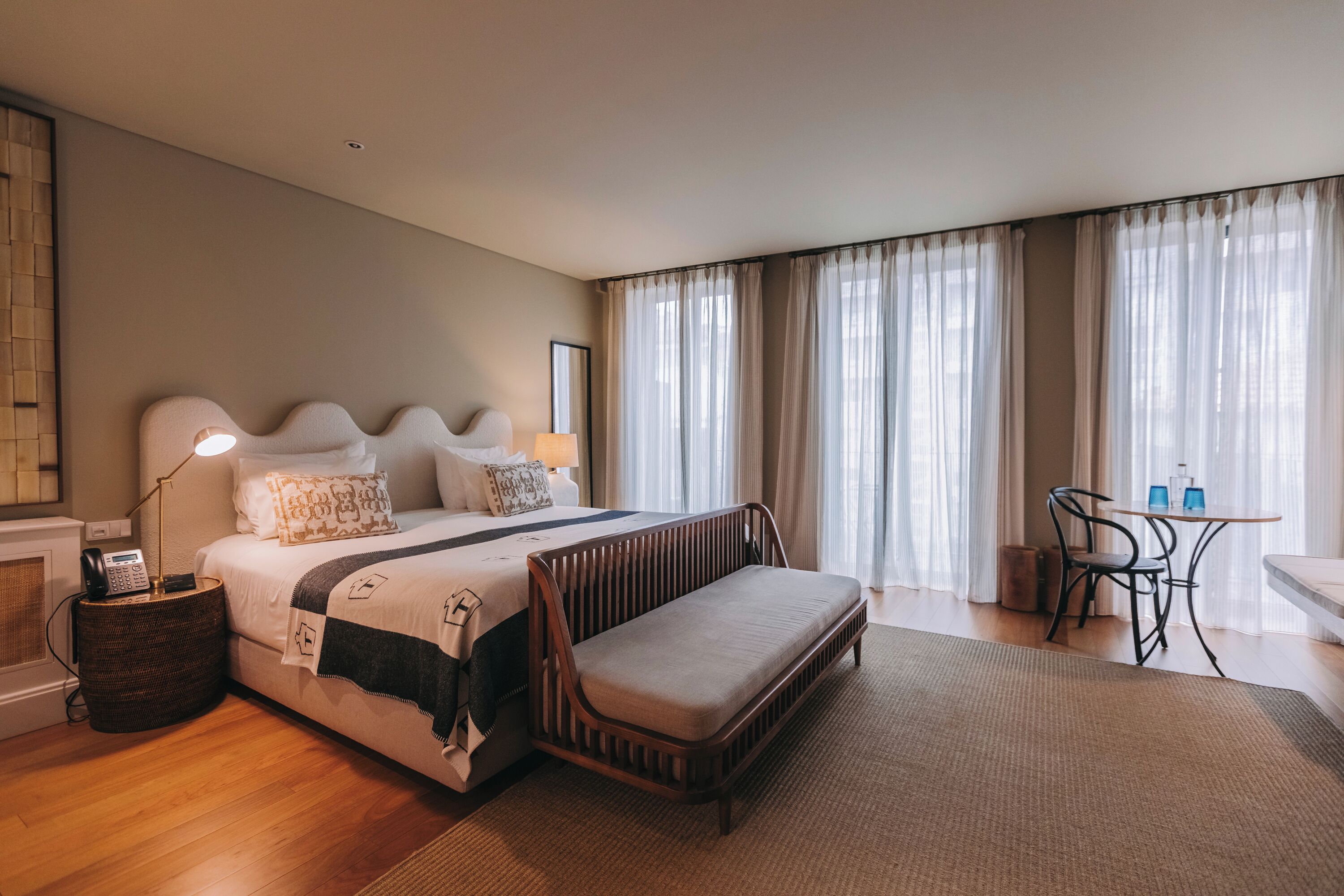 A hotel room with a bed, bedside table, lamp, chair, and a view of the outdoors through large windows.