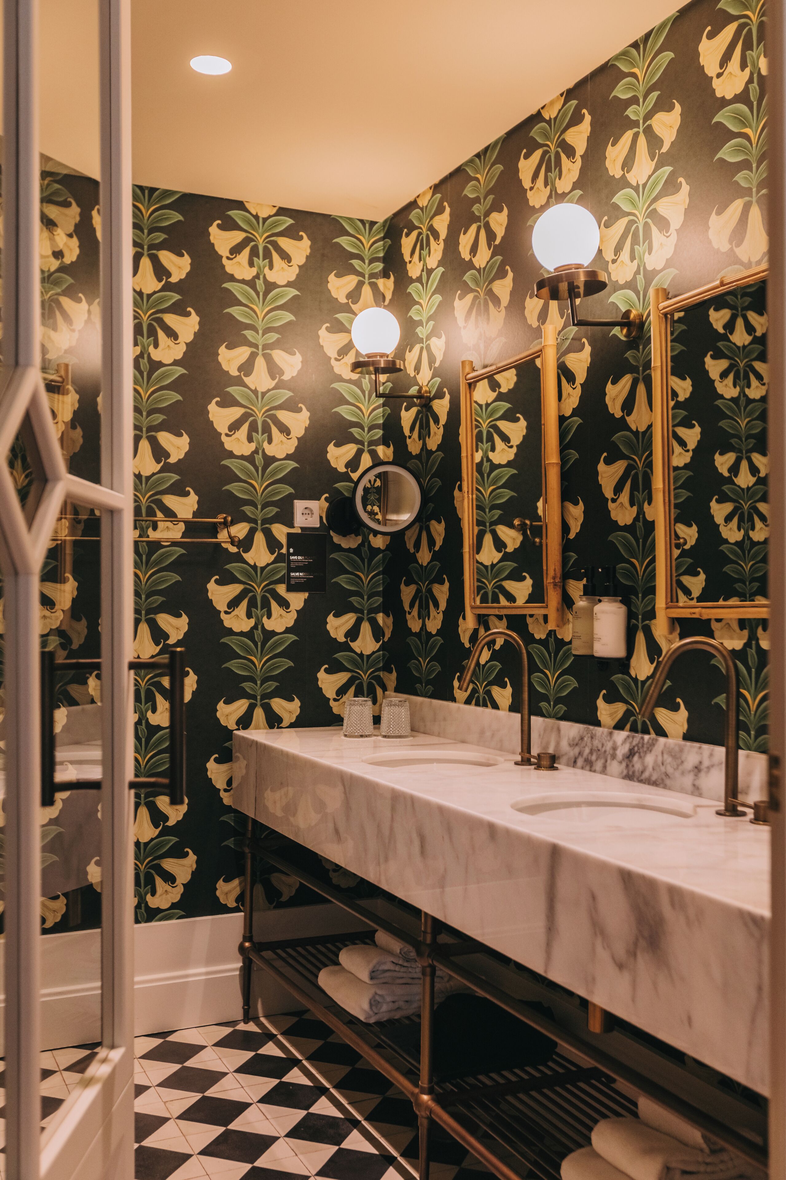 A bathroom with a black and yellow floral wallpaper, a white sink, and a mirror with a round light.