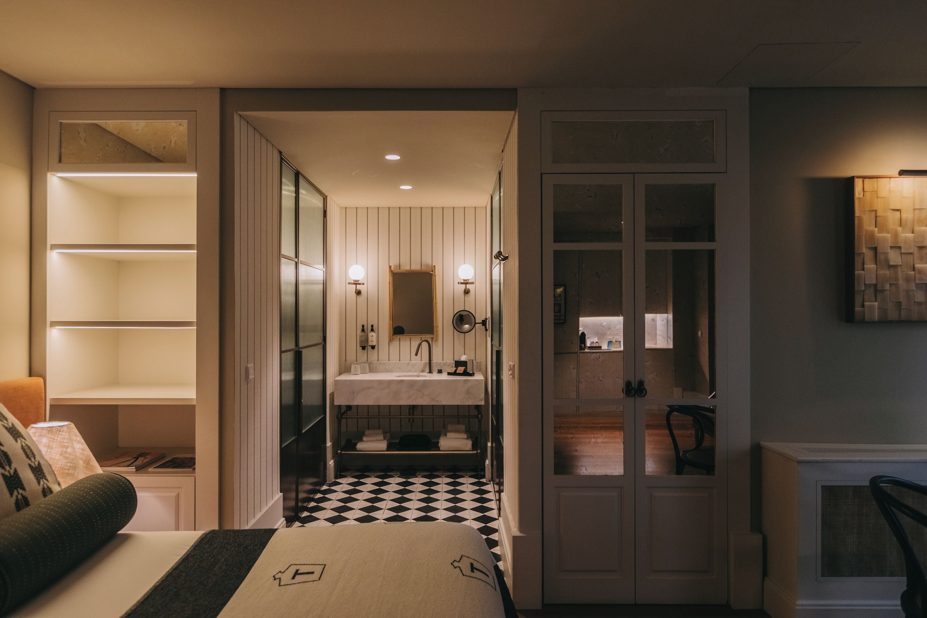 A hotel room with a checkered floor, a bed with a headboard, a bedside table, a lamp, a wardrobe, and a bathroom with a sink and mirror.