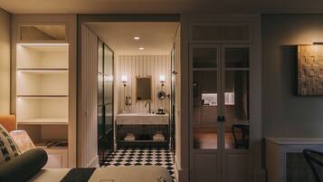 A hotel room with a checkered floor, a bed with a headboard, a bedside table, a lamp, a wardrobe, and a bathroom with a sink and mirror.