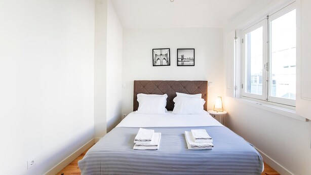 A neatly made bed with two pillows and folded towels, a headboard, a bedside lamp, and two framed pictures on the wall. - Apartamento de 1 quarto em Santa Catarina (Porto)