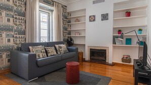 A living room with a grey sofa, patterned cushions, a red stool, a fireplace, and a bookshelf with decorative items.