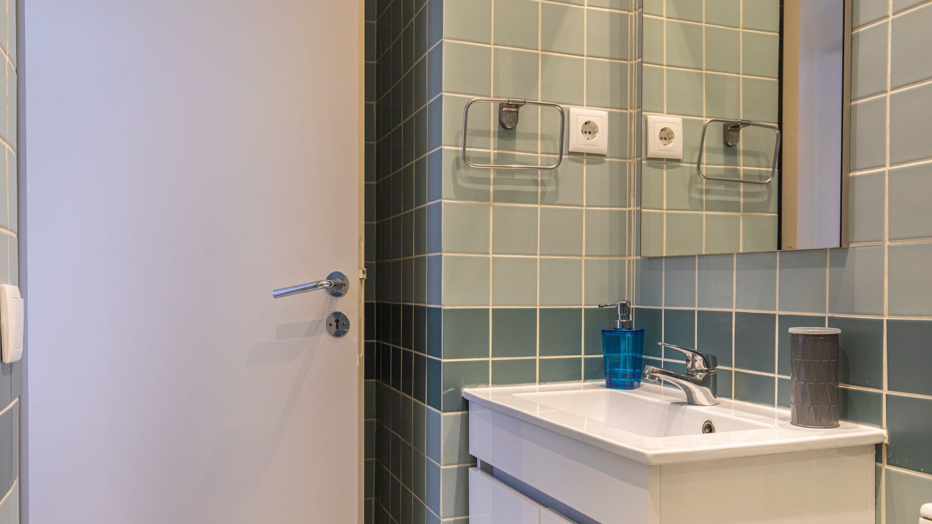 A bathroom with a white sink, a blue soap dispenser, a grey trash can, and a mirror above the sink.