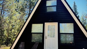 Une cabane triangulaire en bois, avec une porte blanche, deux fenêtres et une véranda équipée de deux chaises.