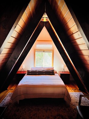 Een slaapkamer met een schuine kap, een bed, een raam en een hanglamp.