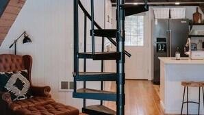 Un loft moderne avec un escalier en colimaçon noir, des planchers en bois, un fauteuil en cuir confortable et une cuisine à l’arrière-plan.