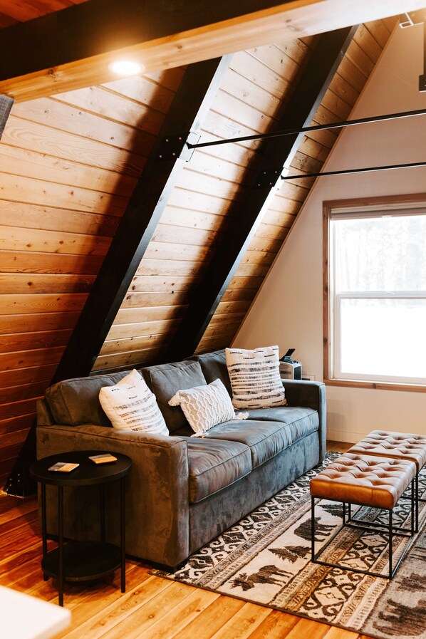 Une pièce de style loft avec un plafond en pente, un canapé gris agrémenté de coussins à motifs, une petite table ronde et un tapis à motifs.