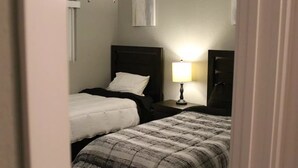 Une petite chambre d’hôtel à la lumière tamisée, avec deux lits simples, une table de chevet avec une lampe et un mur sobre orné d’un seul tableau.