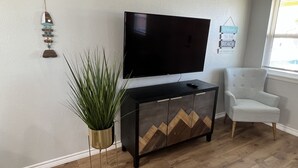 En stue med et fladskærms-tv monteret på væggen, et moderne skab med geometriske mønstre, en potteplante og en hvid lænestol.