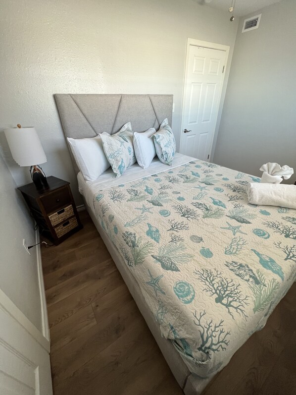 A neatly made bed with a blue and white ocean-themed bedspread, a bedside lamp, and a small wooden nightstand with a lamp.