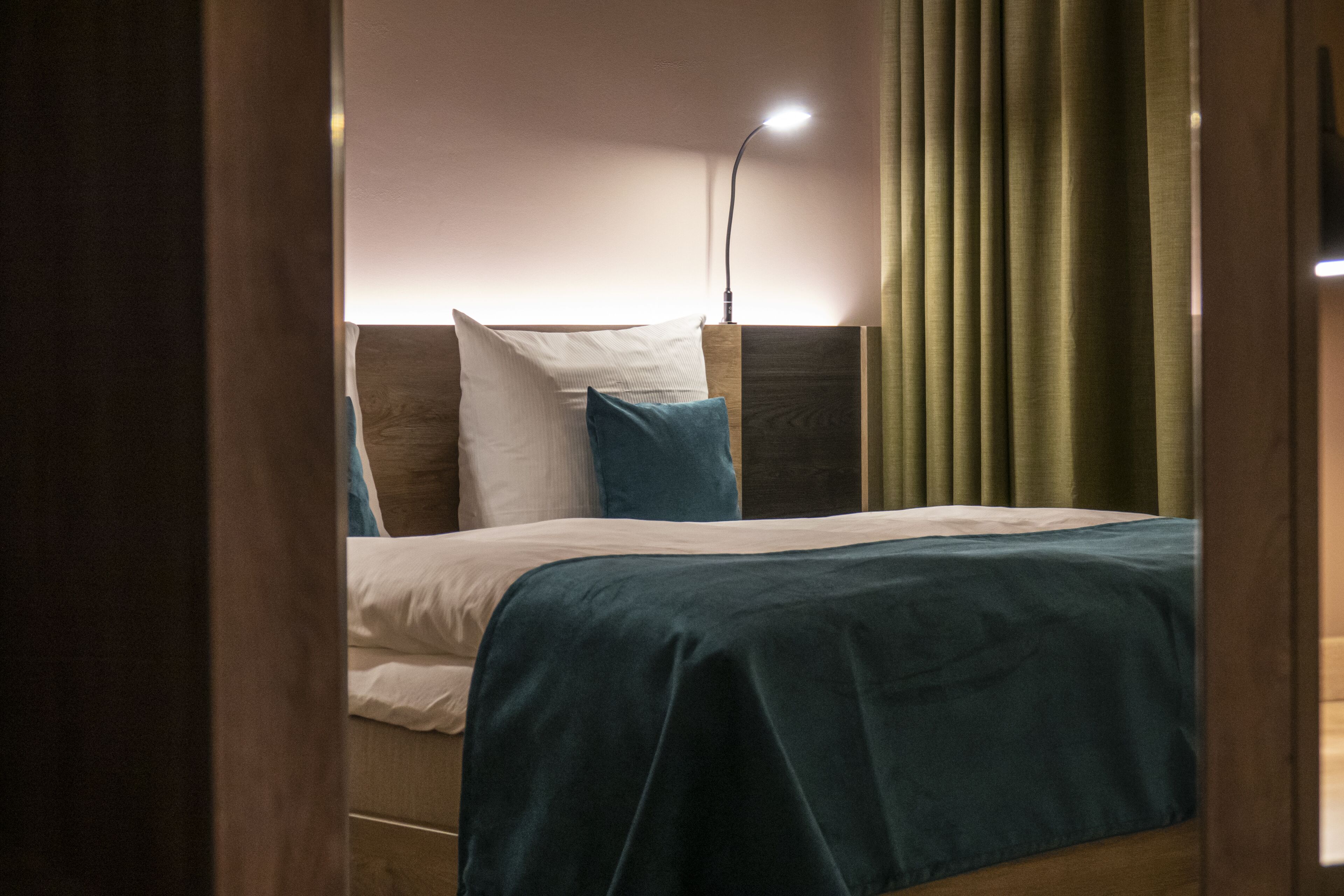 Una cama bien tendida con una colcha color turquesa y almohadas blancas, un cabecero de madera y una lámpara de noche.