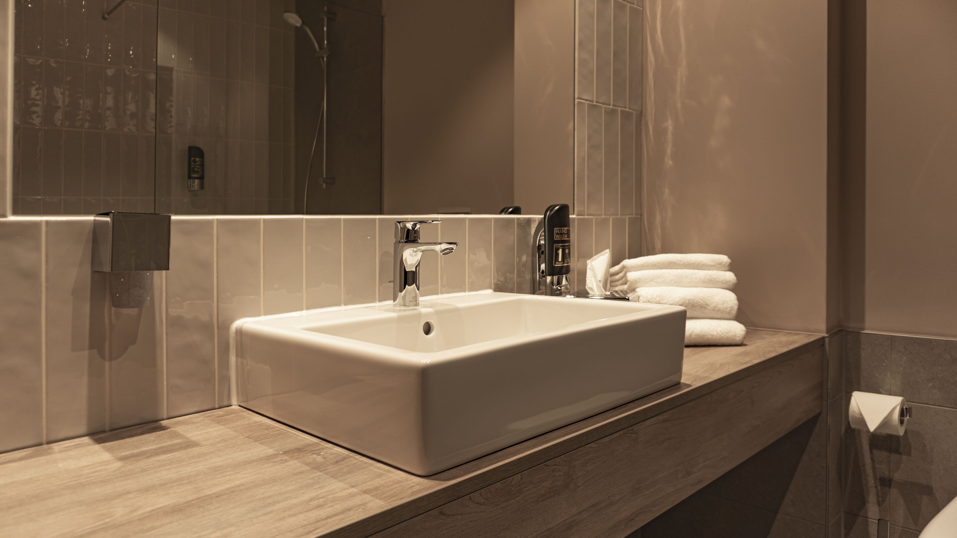 Un baño moderno con un lavabo grande, un dispensador de jabón y una pila de toallas blancas en la encimera.