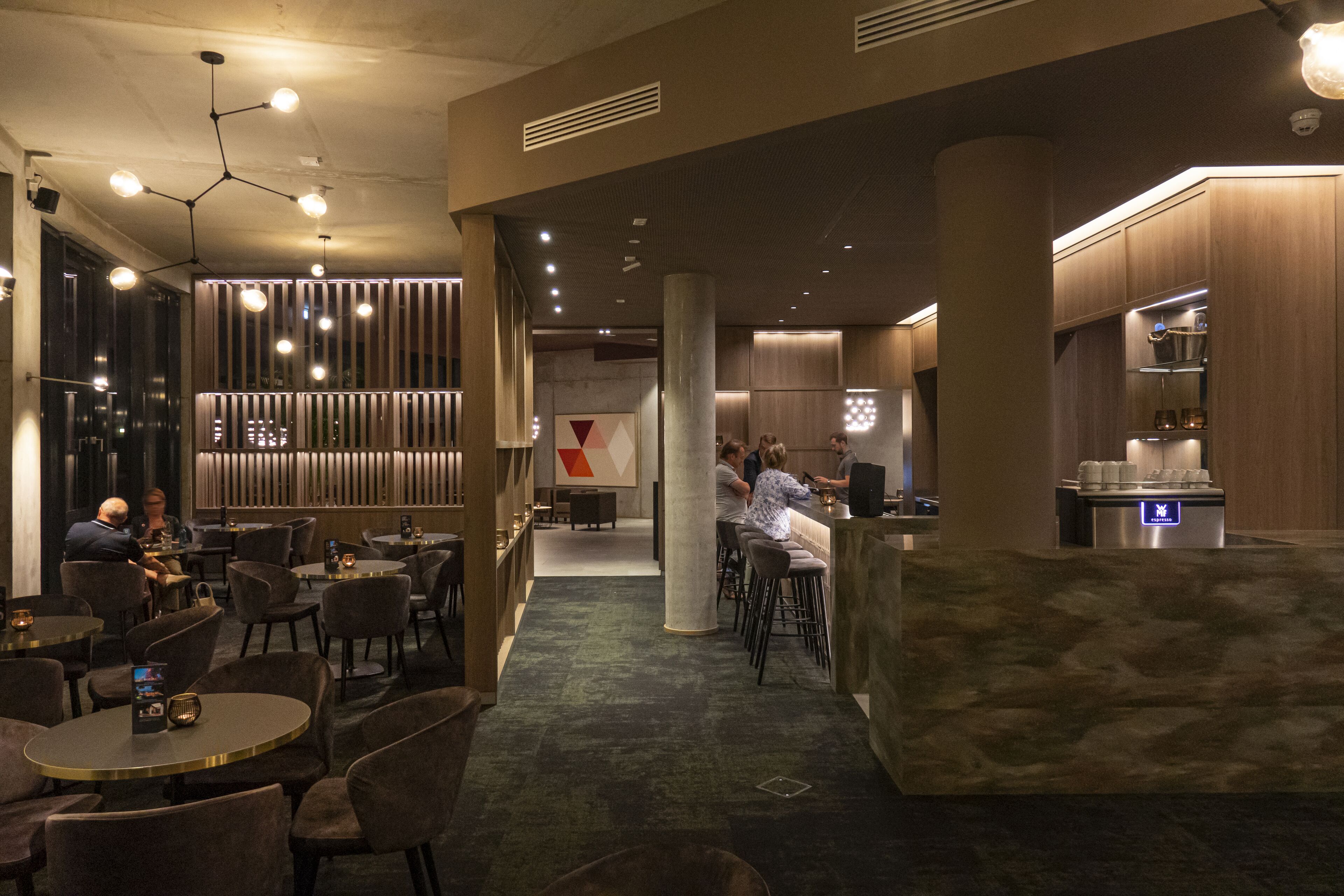 A modern hotel lobby with round tables and chairs, a bar area, and a reception desk.