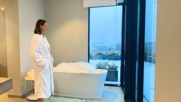 Un baño moderno con una bañera grande, un ventanal con vistas a edificios y una mesita auxiliar con velas.
