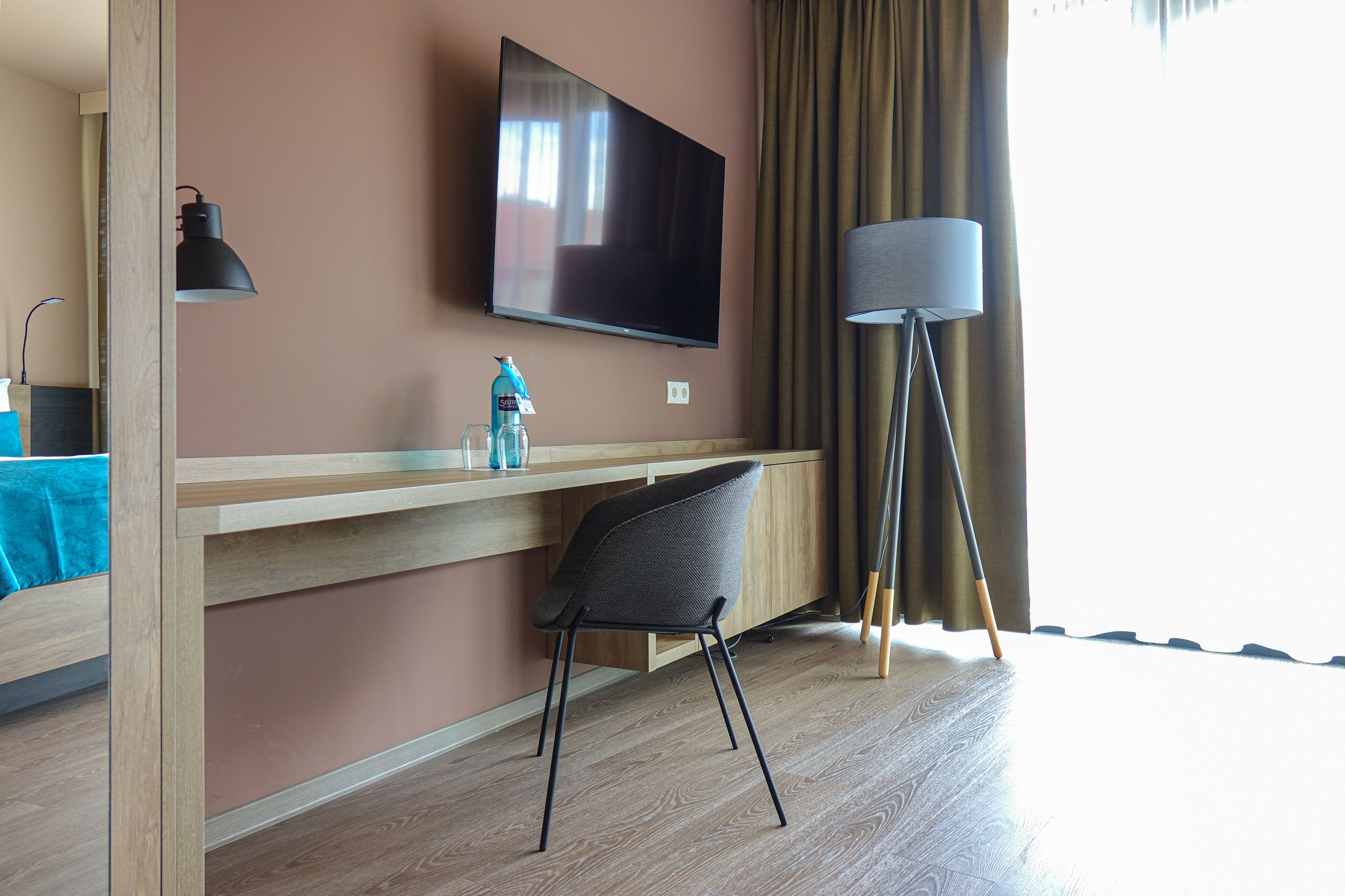 Moderni hotellihuone, jossa on seinälle asennettu taulutelevisio, puinen työpöytä, musta tuoli, lattiavalaisin ja ikkuna, jossa on verhot.