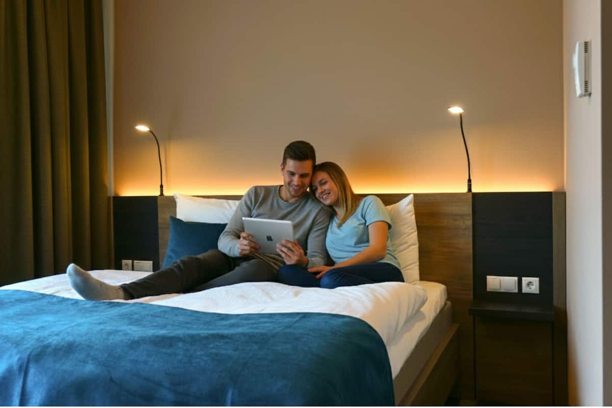 Una pareja sentada en una cama con una tableta, en una habitación de diseño moderno, una mesita de noche y una lámpara.