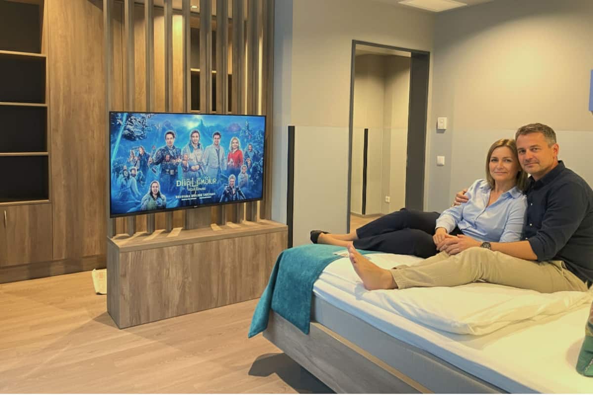 Una habitación de hotel moderna con un televisor de pantalla plana grande montado sobre un soporte de madera, una cama con una manta color aguamarina y un armario de madera.