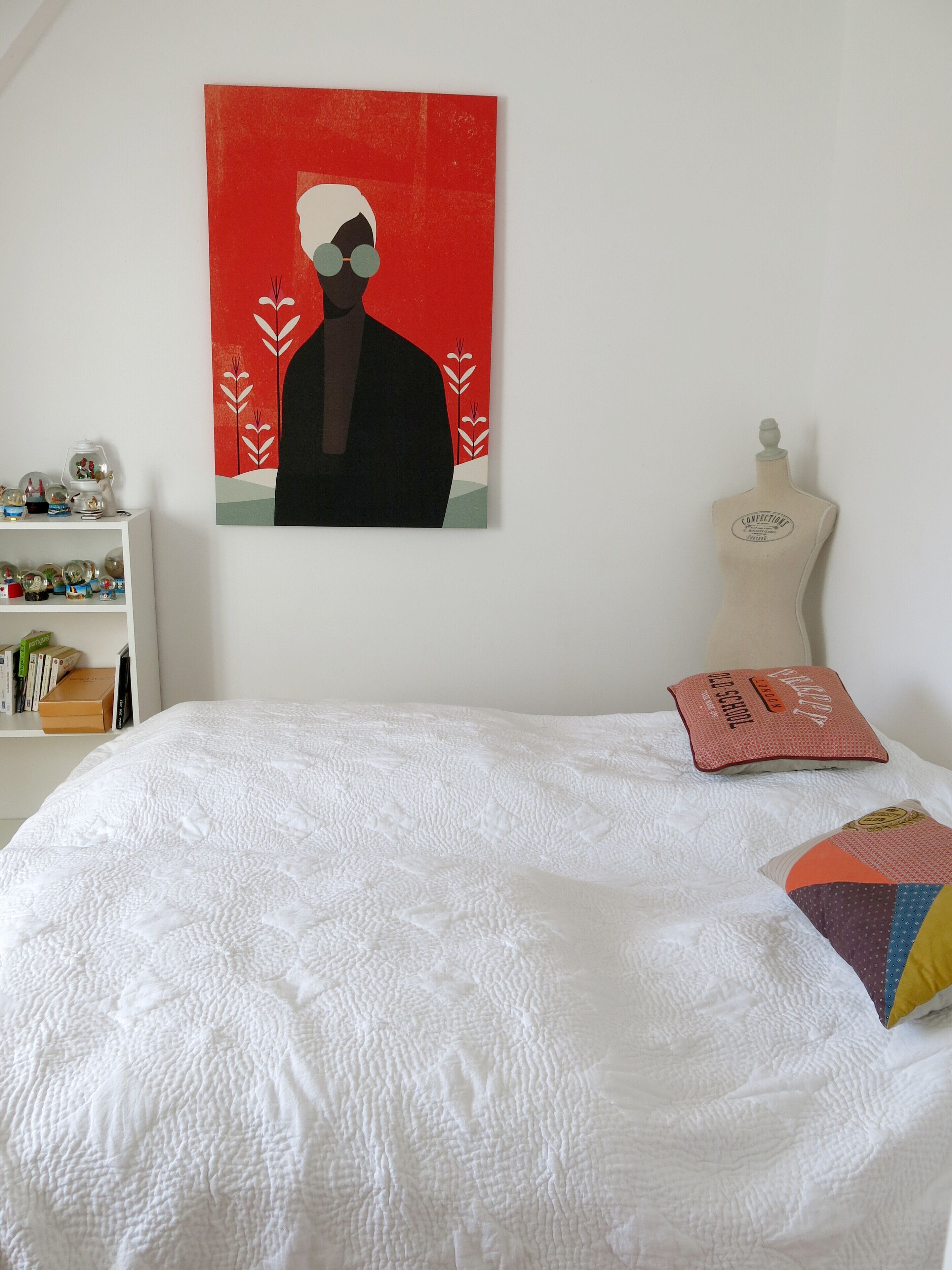 A bedroom with a white quilted bedspread, a colorful pillow, a white dresser with decorative items, and a wall-mounted painting.