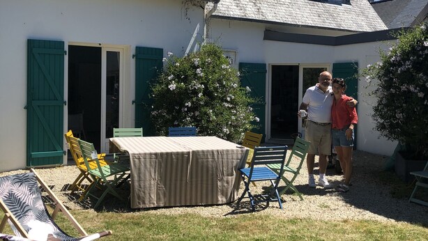 Una casa con techo inclinado, un porche cubierto con mesa y sillas, y un pequeño jardín.