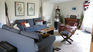Una sala de estar con un sofá gris, una mesa de centro de madera y un sillón rojo. Hay dos cuadros en la pared, una lámpara y una planta alta.