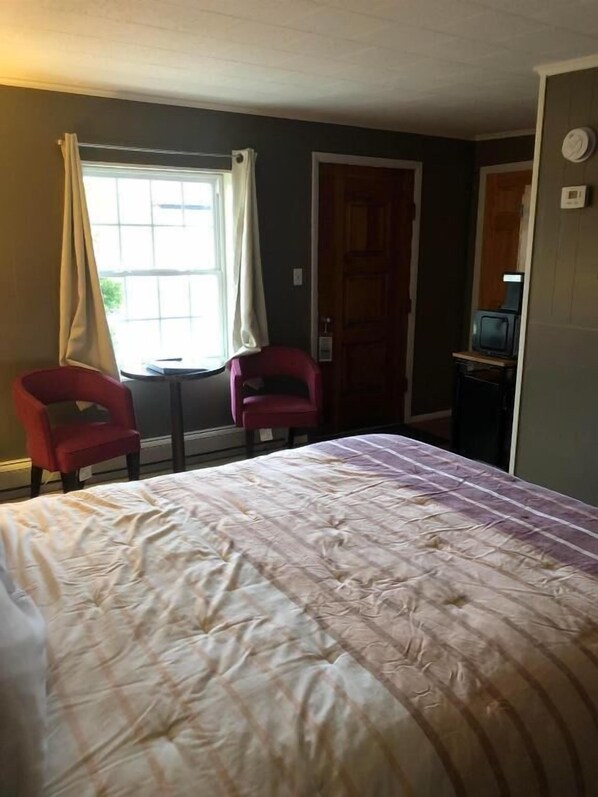A room with a bed, two chairs, a window with curtains, a door, and a small table with a television.