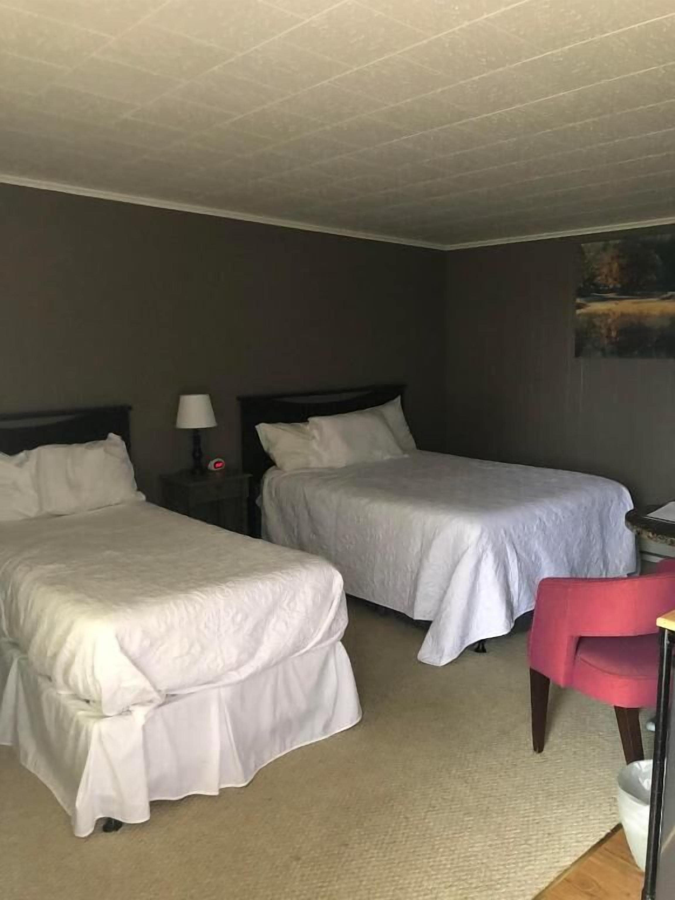 Una habitación de hotel con dos camas, una mesita de noche con una lámpara, una silla roja y un cuadro en la pared.