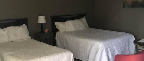 Una habitación de hotel con dos camas, una mesita de noche con una lámpara, una silla roja y un cuadro en la pared.