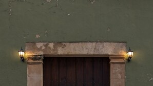 Una puerta de madera oscura con marco de piedra, flanqueada por dos luces fijadas a la pared, sobre un fondo de pared verde.