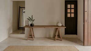 Una habitación minimalista con un banco de madera, una lámpara colgante, una planta en maceta y una puerta oscura con ventanal.
