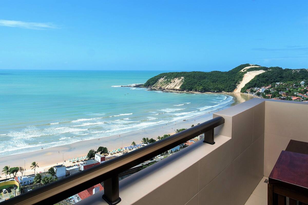 Blick auf einen Strand vom Balkon mit Geländer, strahlend blauer Himmel und ein Sandstrand mit Sonnenschirmen und Gebäuden im Hintergrund.