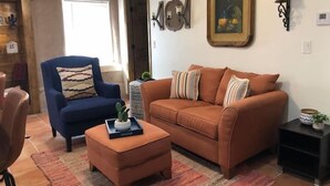 A living room with a blue armchair, an orange sofa, and a small ottoman. There is a painting on the wall and a small table with a plant. - Lovely 1-Bedroom Adobe Escape (Mesilla)