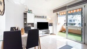 A modern living room with a flat-screen TV, a dining table with chairs, a bookshelf, and a balcony with a view of buildings. - Home2Book Cozy Apartment Los Cristianos, Wifi&Pool (Los Cristianos)