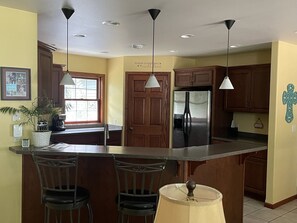 A kitchen with wooden cabinets, a stainless steel refrigerator, a large island with bar stools, pendant lights, and a window with blinds.
