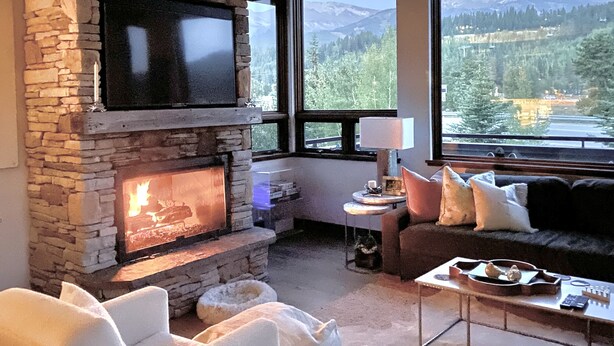 A cozy living room with a stone fireplace, a comfortable sofa, and a view of a lake and mountains through large windows.